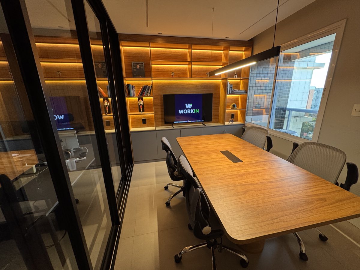 Sala de reunião com mesa de madeira, estante iluminada e o logo da Workin na TV